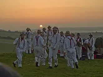 A group of twelve individuals wearing all-white costumes, including hats, are dancing in the centre of a green, grassy space. The low position of the sun indicates that it is early in the morning