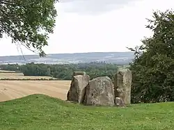 Three large grey boulders stand, adjacent to each other, on an area of grass in the foreground of the picture. In the background, the land drops dramatically to reveal an area of lower farmland.