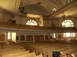 The back of a wide, two to three-story room is visible. Four visible rows of wooden pews lead to the back wall, which has three sets of double doors at its center. A second-floor balcony which projects partway into the sanctuary holds more wooden pews. The back wall has two large arched stained-glass windows visible, and two smaller rectangular ones one each side of the doors. The ceiling is arched, with elaborate chandeliers with Star-of-David shapes hanging from it.
