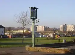 A photograph of an urban park with houses and flats in the distance. The park large, and mostly grass. A football goal is in the centre of the park, and a pub sign is in the foreground.