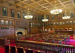 The courtroom seen above, empty but from a different angle showing more of it and its ornate walls, chandeliers and red carpet