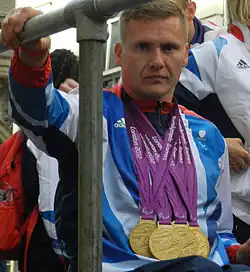 A man in a wheelchair, he wears a white and blue top and around his neck hang four gold medals.