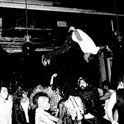A black and white photo of Playboi Carti mid-stage dive, upside down with both middle fingers raised. A dense crowd reaches up toward him in a chaotic concert scene