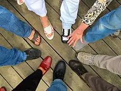 A group of people stood with one foot outstretched, forming a circle. They wear a variety of shoes and sandals.