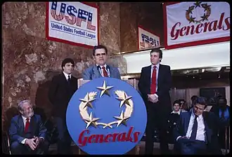 Trump, Doug Flutie, and New Jersey Generals head coach Walt Michaels standing behind a lectern with big, round New Jersey Generals sign, with members of the press seated in the background