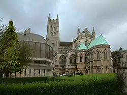 Ornate building with central tower. To the right is a stone building with green roof and to the left a new building with large glass windows.