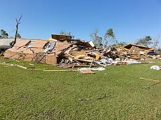 A red brick home surrounded by grass, seen destroyed. A debarked tree stands to its left.