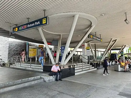 An entrance to the station next to a bus stop, featuring a modern architectural design with a large overhanging white roof supported by distinctive, angled cylindrical columns.