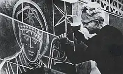 Edward Catich, a white man with glasses and light hair, uses a hammer and chisel to carve an image of a religious figure into a wall