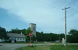 From the right turn lane on Taylor Road, the Bethel Bible Church at left, a rectangular grain elevator building in the center, and Illinois Route 8 sign on the right.