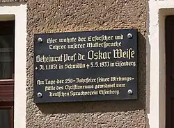 A black sign with gold Gothic lettering on the side of a concrete building