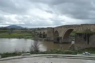 Photo of an old arched stone bridge over a river. There is a mountain in the distance
