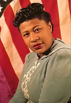 Headshot of Ella Fitzgerald. She was a black woman with dark curly hair. She is a wearing a light green dress with a white flourish in the front. She is standing in front of an American flag.