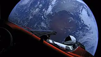 Large circular disc of a fully-illuminated planet Earth showing Australia floating in the blackness of space. In front of Earth is a red convertible sports-car seen from the side. A humanoid figure wearing a white-and-black spacesuit is seated in the driving seat with the right-arm holding the steering wheel, and the left-arm resting on the top of the car door.