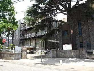 The school building, a large 3-storey building covered with a stone facade and surrounded by low walls.