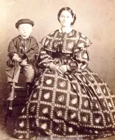 Small blond boy sitting on a stool; to his right a young woman in a full-skirted dress made of fabric with a bold and probably colorful diamond pattern