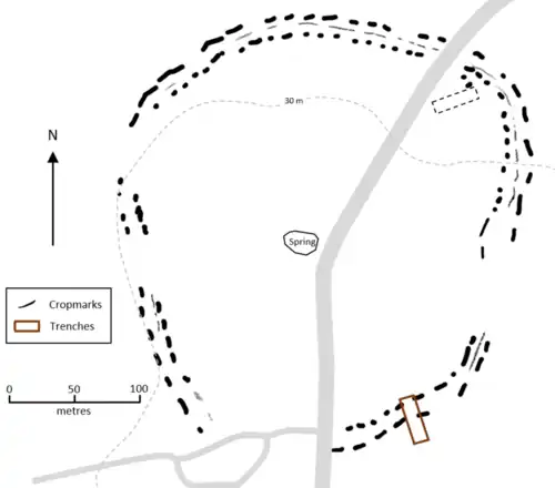 Two concentric more-or-less circular dashed lines, indicating cropmarks, with a thin line occasionally visible between them. A road passes through north to south. A rectangular trench cuts the enclosure line in the southeast corner and there is a small rectangular shape inside the enclosure in the northeast corner.