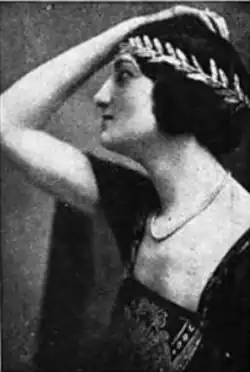 A white woman photographed in profile, with one arm bend and a hand resting on her head; she has dark hair, bobbed and dressed with a laurel-style headband; she is wearing a dark dress with a low wide square neckline