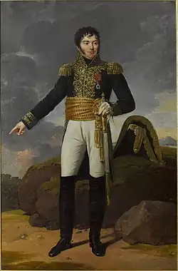 Formal full-length portrait of a Tharreau in uniform, in a rocky landscape. He is a short, solidly built man with a round face and alert dark eyes. His pale skin contrasts with his black curly hair and heavy cheek whiskers. He looks out of the picture to the left while gesturing backward with his right hand. He holds a sword in his left hand.