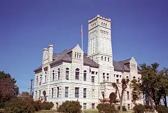 A rough-faced light-colored stone building with pointed roofs and a tower in the center amidst trees and shrubs. An American flag flies from a white pole in the front