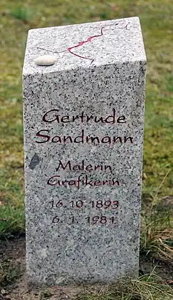 A gray granite headstone reading "Gertrude Sandmann" with grass growing around it.