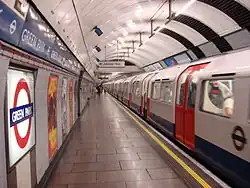 The interior of a building with a rounded, white roof, white walls, grey flooring, and a train on the right carrying passengers