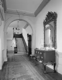 A hallway with rugs stretching through an archway and leading to stairs at end. A console table with drawers sits on the right of the hall with a mirror above it on the wall.