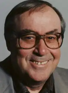 Photo of a middle-aged man with black and grey hair; wearing brown-rimmed eyeglasses, dark plaid dress shirt, and a grey suitjacket