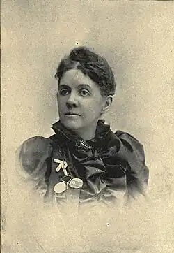 B&W portrait photo of a woman with her hair in an up-do, wearing a dark, flouncy blouse with a brooch, white ribbon, and spectacles pinned onto it.