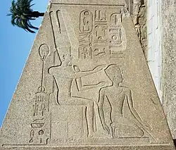 A trapezoidal stone obelisk engraved with Egyptian hieroglyphs and two figures. The figure on the left is seated and wears a tall crown, both hands outstretched to the figure on the right, kneeling, who wears a much smaller crown.