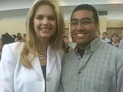 A woman with light skin and blonde hair stands next to a man with dark hair, dark skin, and glasses