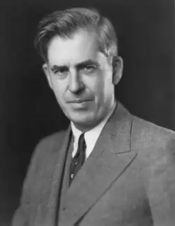 Black-and-white image of the head and shoulders of Wallace with upswept hair, wearing a gray suit and a dark tie