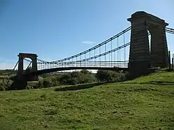 Suspension bridge with stonework arches either end, partly silhouetted against clear blue sky, green field in foreground