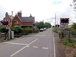 a normal country road passes straight up the middle of the picture, and a two-track railway line crosses it in the foreground. Modern red-and-white barriers protect the track, and are currently up. On the left, just beyond the line, 19th century red brick buildings stand next to the track. This was once the railway station but is now a private house. The line and the road are fringed with hedgerows, those along the road on the left being particularly tall. A field of wheat can just be glimpsed on the right, behind the barriers, lights, and hedgerows.