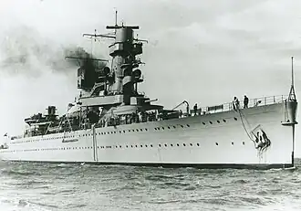 A black-and-white photograph of the cruiser in a calm sea, seen from the bow. Her entire profile is visable, which includes a tall, dominant tower, a large turret on the deck, a smaller turret superimposed near the bridge, and a funnel releasing smoke.