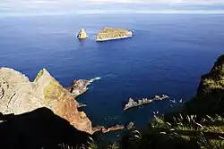 Two small volcanic islands with minimal vegetation as they appear from a neighboring island - the ocean, in dark blue, and the sky, in light blue, are also present in this image