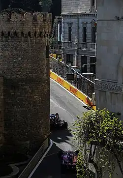 A tarmac race course, lined with concrete and metal barriers, passes between a building and a turret.
