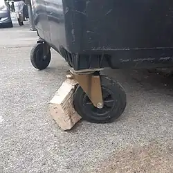 Improvised wooden chock used on a four-wheeled plastic bin
