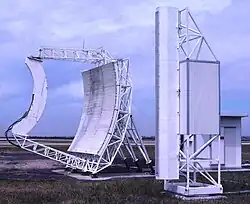 Colour photograph of two large white-painted items of equipment (the left being a parabolic 'dish' with straight sides; the right a tall, narrow structure) on grass, with cloudy sky above.