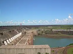 Aerial view of a huge concrete structure located between some low hills and a river, with some large pipes at the base and power lines at the top of the structure and a road