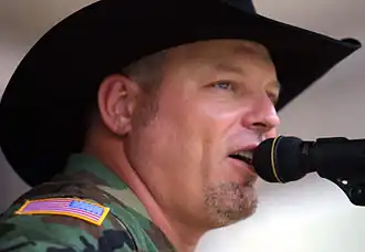 A head shot of John Michael Montgomery, wearing a black cowboy hat and singing into a microphone