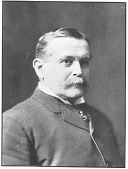 A bust-length black and white photo of a man in a tight collared jacket with short hair and a bushy mustache