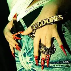 Close-up on female hands with nails done sharp, resting on denim covering legs, blinged out chain reading "Wild Ones" and underwear reading "Jessie Murph".