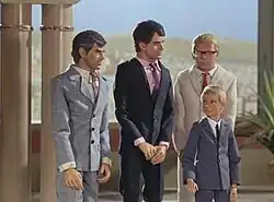 Three men and a boy stand in a palatial setting. A desert landscape is visible from a balcony in the background. One man on the far left is grey-haired and wears a grey suit and tie, the man to the right of him dark-haired and in deep navy blue. Both men are orientated in the direction of the (blond-haired) boy, who is also formally attired in grey. The third man, also blond but wearing a cream-shaded suit, stands directly behind him.