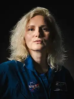 A woman in a blue flight suit is standing in front of dark background; she is facing the camera with a slight smile.