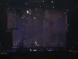 A stage with a big screen display showing a man with a beard. Sitting in a piano, a man with long curly hair wears a white shirt, with the sleeves roll back, and a black vest, plays the piano. To the man's left, a man can be seen standing playing an electric piano.