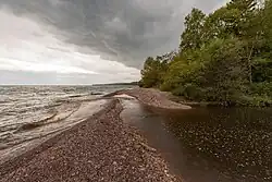 A stream of dark water flows from the right side of the images, away from the camera and some ways down a stony reddish beach, past a copse of green trees, before veering left across the beach and meeting a lake so wide that its far shores cannot be seen. The sky is cloudier on the right side, less so on left over the lake.