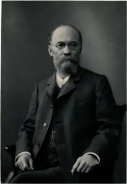 A balding man with round glasses and a long beard wearing a suit looking off camera