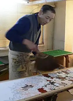 A man hand-dyes a length of white cloth with a floral design, painting in individual flowers in different colours.
