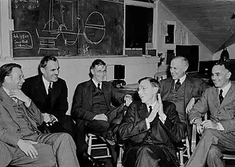 Six men in suits sitting on chairs, smiling and laughing